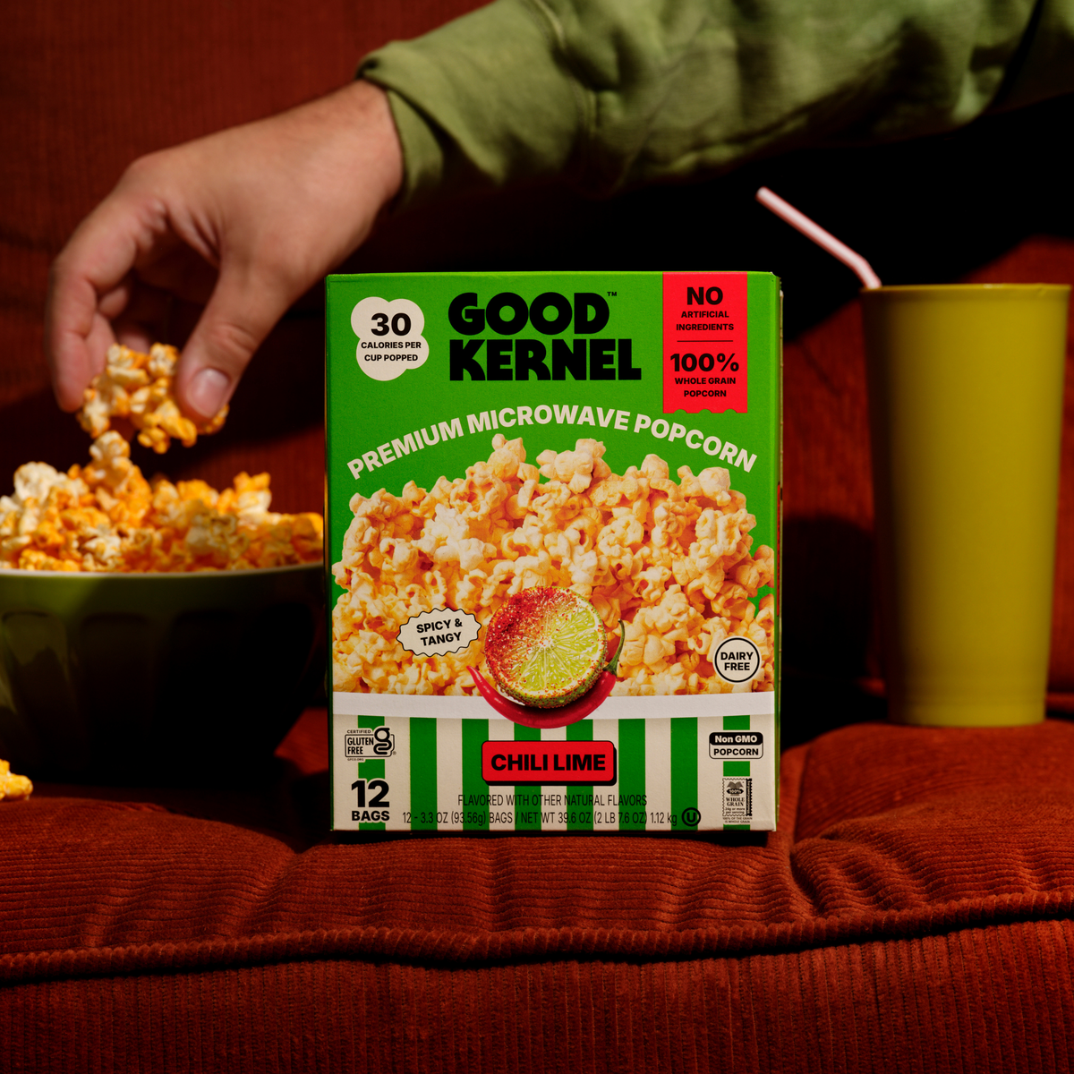 Hand grabbing Chili Lime popcorn next to the Good Kernel box on a couch