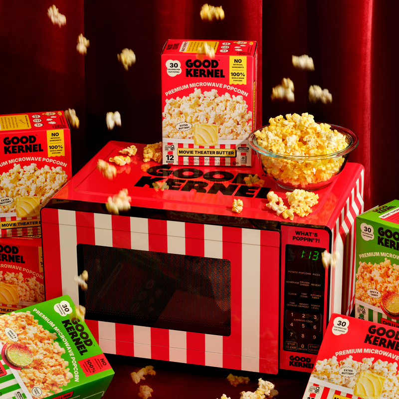 Good Kernel popcorn boxes around a striped microwave with popcorn flying out