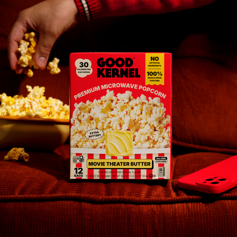 Hand grabbing Movie Theater Butter popcorn next to the Good Kernel box on a couch